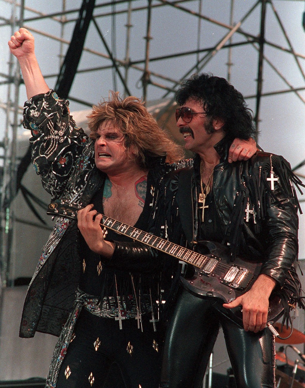 Ozzy Osbourne performing on stage with Black Sabbath guitarist Tony Iommi, both dressed in black leather with cross-themed outfits during a live concert.